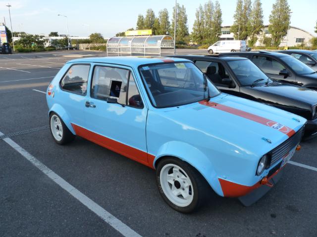 Present Pistonheads - The image shows a bright blue and orange Fiat car parked in a parking lot. Several other cars and vans are also visible in the background, indicating the car is part of a group or parked in a commonly frequented area. There's a clear sky and it appears to be daytime, contributing to the vibrant colors of the Fiat. The car has a poignant expression courtesy of one of the stickers on the back, reading "Still in my feelz," which plays on a meme/phrase often used to describe the feeling of how someone looks or behaves.