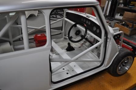 Race Engine builder in the midlands - Page 1 - Classic Minis - PistonHeads - The image shows the interior of a compact race car that is white in color. Inside the vehicle, a black steering wheel is situated in the center. The steering wheel is accompanied by other necessary components for the car's operation, although they are not specifically identifiable. The car is parked in what appears to be a workshop, as evidenced by the visible cardboard box and additional car parts in the background. The texture of the wooden table contrasts with the metallic finish of the car.