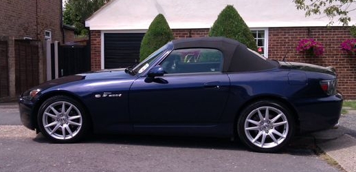 S2000 for 1980's 911?  Ummm....er....well.... - Page 1 - Porsche General - PistonHeads - The image shows a dark blue convertible sports car parked on an asphalt surface. It appears to be a daytime setting, with taller brick buildings in the background. The car model is indicated as a Mazda Miata, as suggested by the badge and design features visible on the vehicle. The car is convertible, with a black soft top roof retracted, and it's a two-door model with the doors closed.