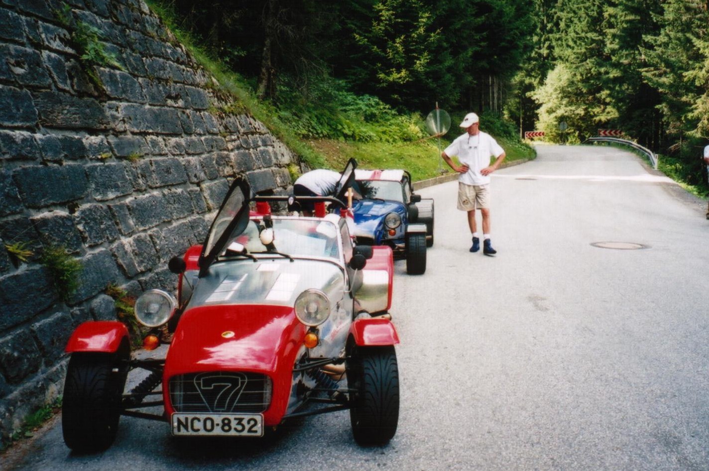 RE: PH does the Alps - Page 2 - General Gassing - PistonHeads - This image captures a unique sight on a road flanked by a stone wall. Two small, brightly colored cars dominate the scene; one is a vibrant red with a prominent white stripe running down the middle, the other is similar in color scheme but in shades of blue. They appear to be open-topped convertibles, adding to their charm. A man, seemingly enjoying the scene, stands on the road next to the red car. He is dressed in a white cap and a white shirt, providing a stark contrast against the colorful backdrop. 

He is positioned near the stone wall, which extends into the background. Beyond the wall, the lush greenery of trees can be seen, suggesting that this could be a rural or semi-rural area. The image exudes a sense of leisurely exploration or tourism.