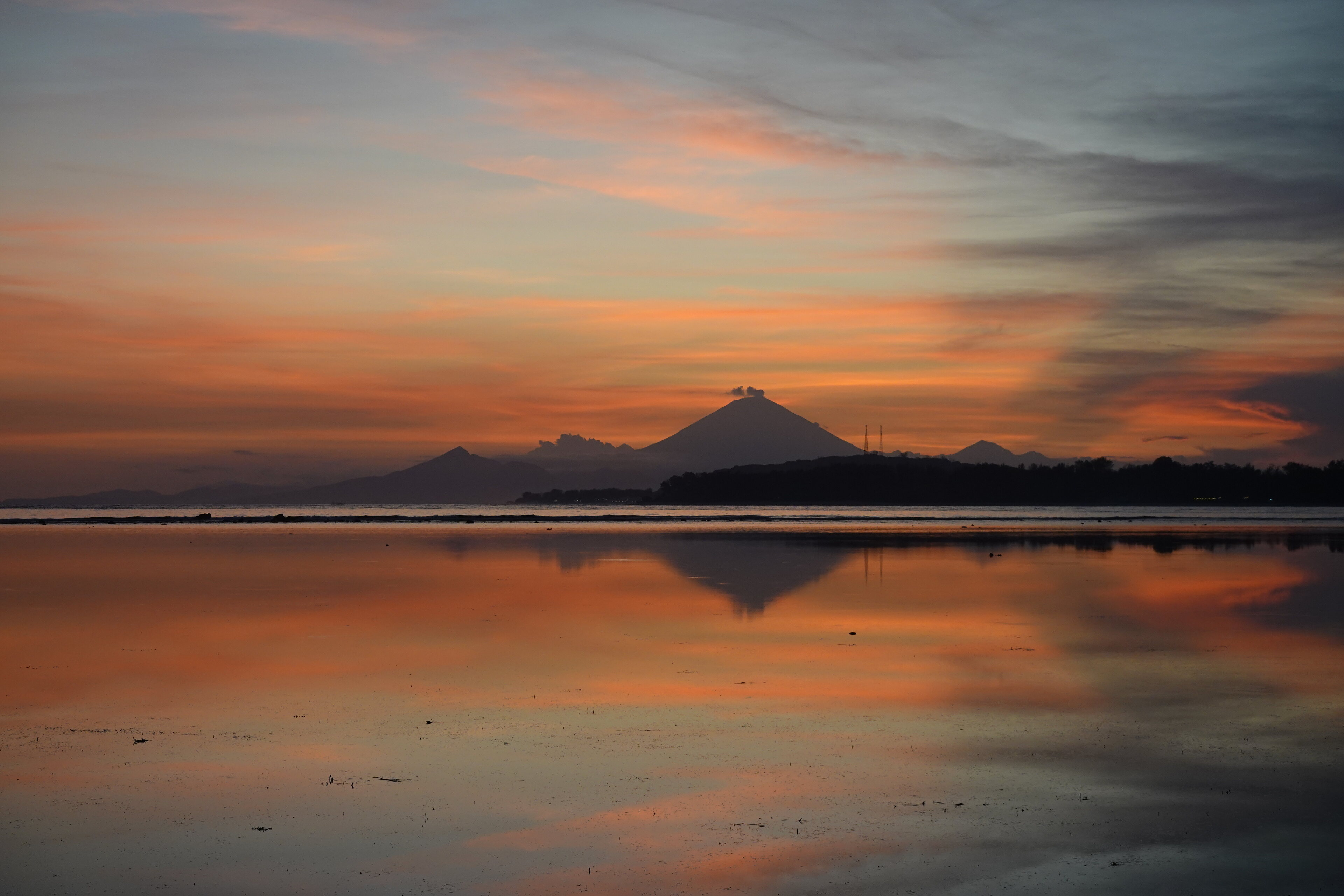 Pistonheads - The image captures a serene scene of a sunset at a beach. The sky is painted with hues of orange, pink, and purple, reflecting on the calm water below. In the center of the image, there's a small island or mountain peak rising majestically from the horizon. The foreground of the image showcases a wet sandy beach, which appears to be tidal, as evidenced by the tide marks on the right side. The overall atmosphere is one of tranquility and natural beauty.