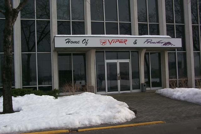 A red brick building with a sign on it - Pistonheads - The image shows the front entrance of a building, which has white pillars and large windows. Above the entrance, the words "Home Of" are visible, and just underneath that, there are the names "VIPER & FUELTAMPER" written in a bold, colorful font. In front of the building stands a path with snow on either side, leading up to the entrance where a door is partially open. To the right of the entrance, a garden with bare bushes adds to the wintry ambiance.