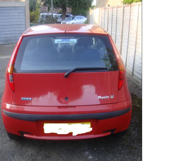 Pistonheads - The image shows the rear view of a small red car parked at an angle on an asphalt surface. A metal fence can be seen behind the car, and the gate of a house is partially visible on the left side of the photo. The vehicle appears to be a hatchback model, and the license plate is clearly displayed. The car has a glossy finish and seems to be in good condition.