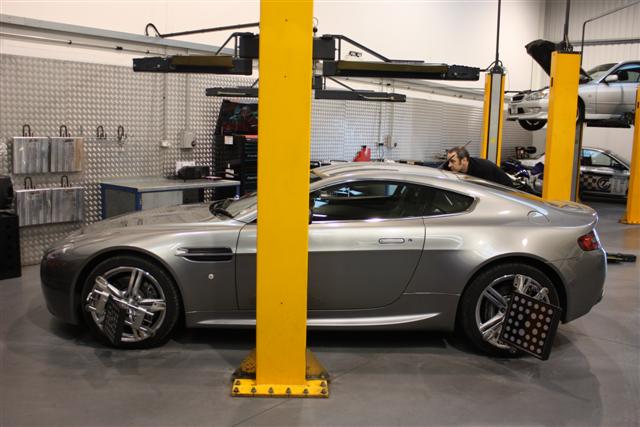 V8 Vantage tuning (SLINC) - TDI Inspection Service - Page 1 - Aston Martin - PistonHeads - The image shows a car lifted on a hydraulic lift with its front wheels raised, indicating it is most likely parked in a workshop or garage. A man is working on the car, attending to tasks under the hood, which is open. The car appears to be a convertible, suggested by the absence of a roof and its open top. The car's color scheme consists of a combination of chrome and non-chrome components. The interior is not clearly visible. The setting suggests a professional environment dedicated to the maintenance and repair of automobiles.