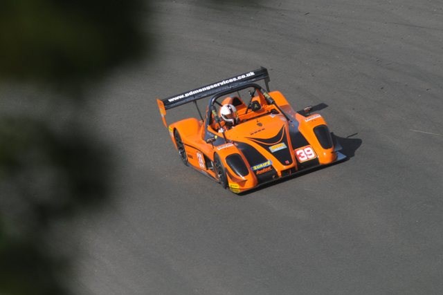 Juno Is Nob Less - Page 2 - Noble - PistonHeads - The image captures a dynamic scene on a race track. In the foreground, an orange and black race car is seen in motion, its sleek body streamlined for speed. The driver, clad in a white helmet and suit, is inside, focused on the track ahead. The race car is equipped with a large rear spoiler and a distinctive black splitter, enhancing the car's aerodynamics. The background is a blur of green, suggesting a well-manicured track, and the anticipation of a high-speed chase is palpable, creating a sense of thrill and excitement.