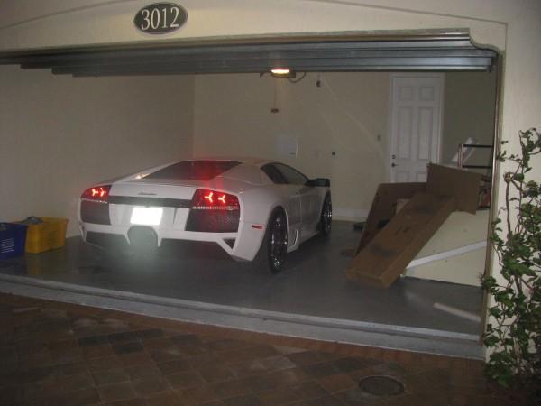 Who has the best Garage on Pistonheads???? - Page 44 - General Gassing - PistonHeads - The image shows a white high-performance car parked inside a garage. The car features a distinctive rear spoiler and a license plate with the numbers "3012". To the right of the car, there is some cardboard debris, indicating recent activity in the vicinity, possibly related to the car. The garage has an automatic door that is currently open, revealing an exterior street view in the background. There is a small amount of foliage to the right side of the garage, likely from a nearby plant. The overall scene appears to be a snapshot of a typical, albeit luxurious, garage space.