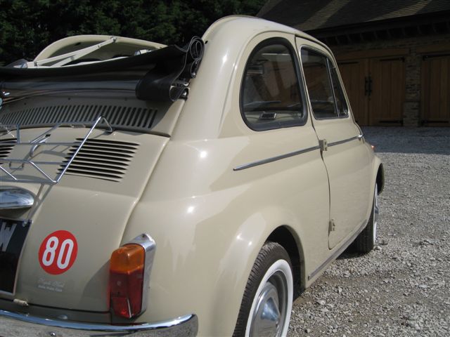 Fiat 500 - Page 1 - Readers' Cars - PistonHeads - The image shows a vintage car parked on a gravel surface with a greenish hue suggesting a natural setting. The car is tan in color with a vibrant, clear red taillight that stands out against its body. The vehicle has an official emblem with a number 80 on it, and its rectangular windows reveal the interior as dark. In the background, sloping, wooden buildings under a grey sky form a rustic atmosphere.