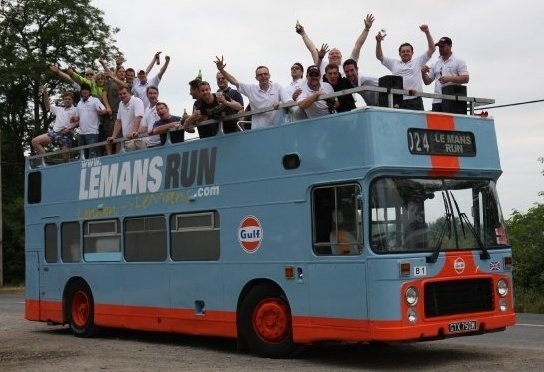 Hrs Lemans Pistonheads - The image features a large, blue double-decker bus parked on a road. The bus is positioned so that its rear is facing the camera, and its windows and sides are adorned with various decorative elements. There are numerous people standing on top of the bus, giving a sense of height and excitement. The text "LEMANS RUN" is prominently displayed on the bus, along with other inscriptions like "GUILG". The bus has a prominent rear buffer, and the color scheme is mostly blue, accented with red on the bottom and orange highlights on the top. The scene seems to be set in an outdoor area, possibly during the day, and the road is clear, suggesting the bus might be on display or awaiting an event.