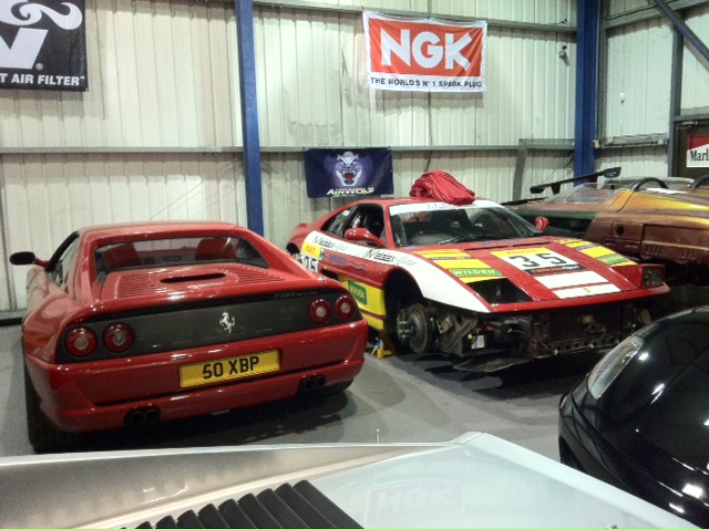 The GT Cafe Gallery - post your meeting photos here - Page 1 - North West - PistonHeads - The image depicts an indoor space, possibly a garage or a warehouse, where several sports cars are showcased. Two prominent sports cars are a red Vauxhall Nova with "60 XB" and "50 XCP" text on the side, and a red and white racing car with the number "46." In the background, a banner with logos of sponsors flanks the room, suggesting the presence of an event associated with the cars. The setting is characterized by a sparse storage arrangement, with a high ceilings and corrugated metal walls.