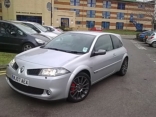 Any PH'ers in the Bridgwater/Burnham/Glastonbury area? - Page 2 - South West - PistonHeads - The image features a silver Renault car parked in a parking lot. The car is positioned at the front of the frame, and it prominently displays the license plate "J70 AEA". Surrounding the car are several other vehicles, including a black, a white, and a blue car. In the background, there is a charming brick building, a small group of people, and a solitary fire hydrant. The overall scene suggests a normal day at a public parking area.