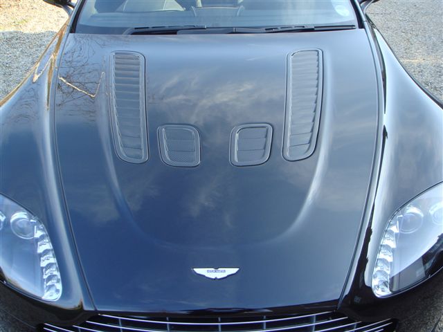 New Vantage V12 - Page 1 - Aston Martin - PistonHeads - The image shows the front of a metallic-blue luxury vehicle. The car is positioned at an angle that provides a view of the hood and part of the nose cones, suggesting that the photo may have been taken during daylight hours due to the lighting. The design of the hood and the ventilation system are noticeable, with two black vents above the grille. The vehicle is adorned with the Aston Martin logo, which is located on the upper part of the grille. The overall style of the image is a close-up, realistic photograph that captures the sleek exterior of the car.