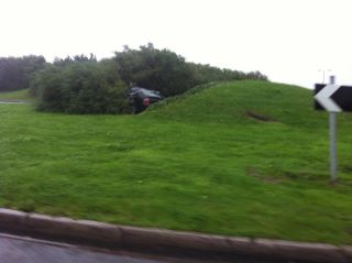 Rover and out! Cribbs crash. - Page 1 - South West - PistonHeads - The image shows a grassy area with a car that appears to have been abandoned. The car is partially buried in the grass and under bushes, suggesting it has been there for an extended period. There is a clear, unobstructed view of the car from the perspective of the road. A street sign with an arrow, typically used for direction, is visible at the bottom of the image, indicating the direction of traffic flow. The overall scene communicates a narrative of disuse and the passage of time due to injury or mechanical failure.