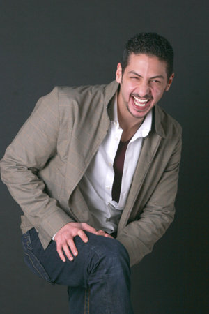 In the image, a man is captured in a moment of joy. He's dressed smartly in a beige jacket and white shirt, adding a touch of elegance to his pose. His arms are wrapped around his thighs and he's knocking his knees against them, a playful gesture that suggests he's having a good time. The backdrop is plain black, putting all the focus on the man's lively demeanor.