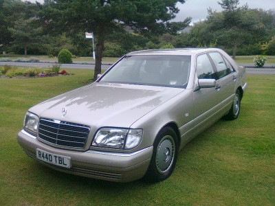 Best Smoker Barges - 1 - 5 Large [Vol 2] - Page 5 - General Gassing - PistonHeads - The image shows a light-colored Mercedes-Benz car parked on what seems to be a grassy area, possibly in a park or a landscaped garden. It is a daytime shot, and the car is facing towards the left side of the image. The weather appears to be overcast or dreary, which gives the photo a somewhat muted color palette. There is a small tree with a visible trunk partially on the left side of the image, and a partial view of a bush is visible to the left of the car. There are no people or other vehicles visible in the image.