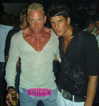 Scott Alexander - Page 91 - The Office - PistonHeads - The image shows two young men, presumably at a party or nightclub, standing together and taking a photo. The man on the left appears sweaty, wearing a white top elevated slightly above the waist, and jeans with a visible loop and belt. The man on the right is dressed in a black shirt with intricate designs on it and matching black pants. Both are making intense expressions and seem engaged in conversation or a shared secret. The background of the image is blurry, focusing attention on the two central figures.