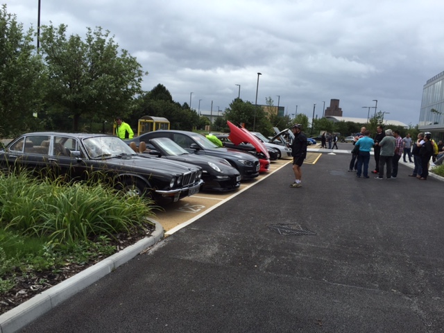 Wirral meet 10.07.2016 - Page 1 - North West - PistonHeads - The image depicts a scene in a large, open parking lot. The lot is filled with a variety of cars, including a black car parked directly in front of the camera. A person is walking towards the right side of the image, carrying a red object that appears to be a water bottle. There are also two people standing to the left of the image, one of whom is wearing a yellow jacket. In the middle and right of the frame, there are trees, providing some green contrast to the concrete and metal of the cars and street light.