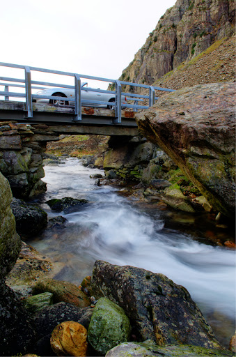 Why are there so few car photographs? - Page 10 - Photography & Video - PistonHeads - The image captures a moment of serenity in a natural setting. The picture features a narrow, metal bridge spanning a gentle river that lightly flows between rocks and stones. The bridge is simple, with a railing on one side, providing a safe passage over the water. Beyond the bridge, a steep mountainous terrain rises, adding a sense of grandeur to the scene. The lighting suggests it might be dawn or dusk, pointing to the time of day when the natural world is most tranquil. The image is a testament to the beauty of untamed nature, untouched by urban development.