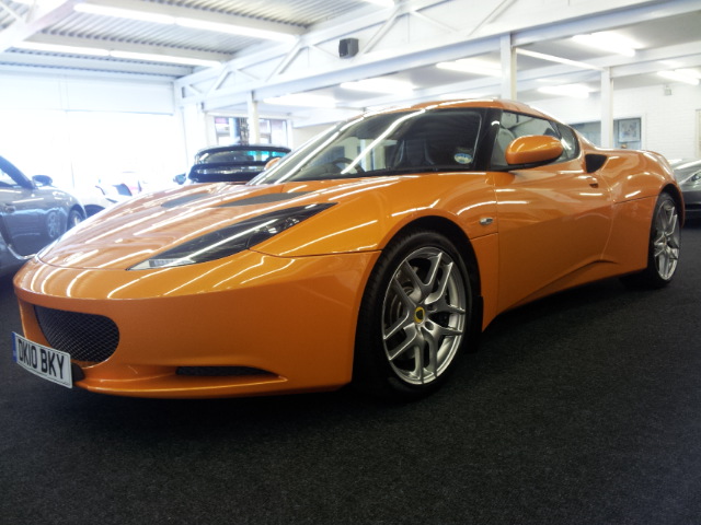 The future is bright.......... - Page 1 - Evora - PistonHeads - The image shows an orange sports car parked indoors. The car's sleek design and curved lines suggest a coupe body style with a fabric roof. It features a sloping front with sharp headlights and a distinctive front grille. The car has five-spoke wheels and a visible registration plate. The indoors setting is illuminated with artificial light.