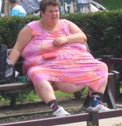 A man sitting on a bench in a park - Pistonheads - The image depicts a woman seated outdoors on a wooden bench. She is wearing a vibrant, floral sundress and has various items around her, including a handbag and a pair of shoes. The woman appears to be engaged in an activity on her phone, possibly browsing or texting, as evident from the device she's holding. The surroundings include a neat hedge and a lane with traffic, suggesting an urban setting. The day seems to be pleasant and clear.