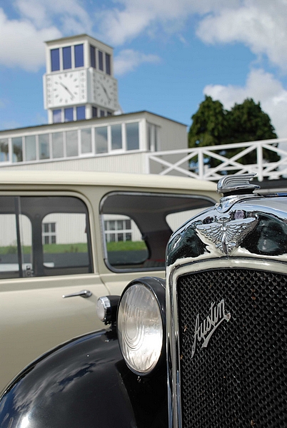 Goodwood Breakfast Club, Pre 1966 classic sunday - Page 1 - Goodwood Events - PistonHeads - The image showcases a vintage Austin car with an antique buff bot style grille. The car is parked in front of a white building that features a white clock tower at one corner. The surroundings indicate a cloudy day. The grille of the car displays the manufacturer's name, and a small headlight contributes to the aesthetics of this classic vehicle.