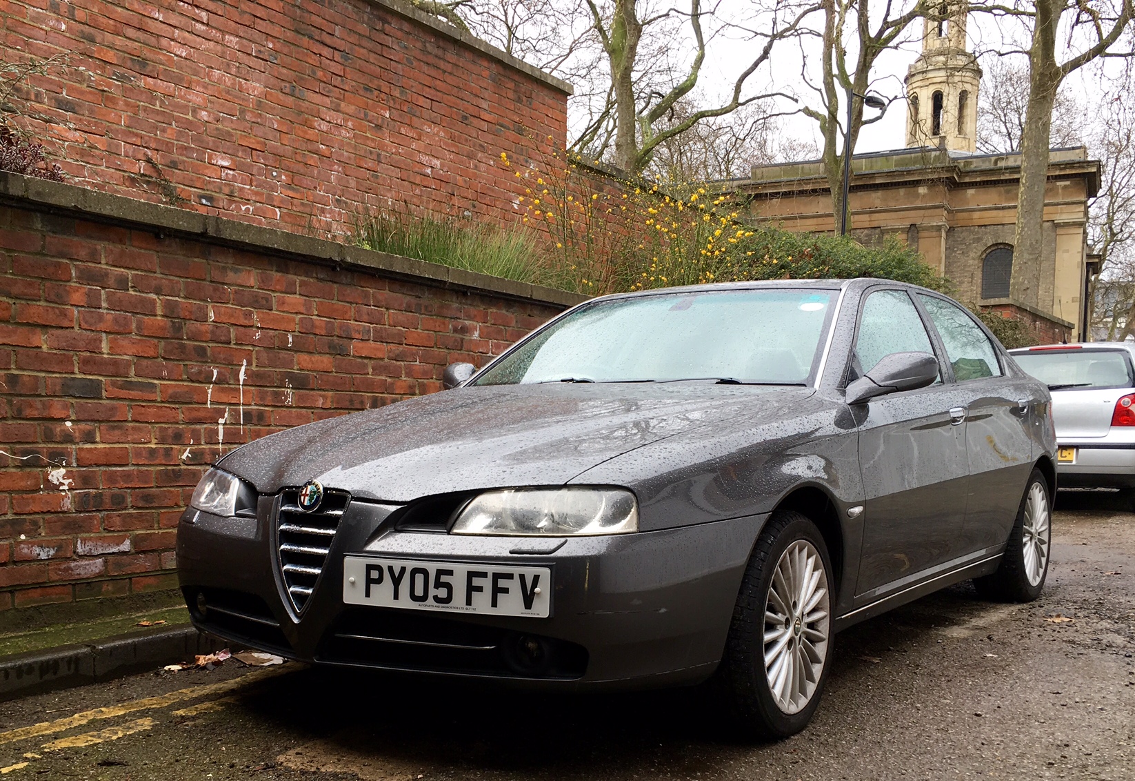 RE: Shed of the Week: Alfa Romeo 166 - Page 4 - General Gassing - PistonHeads - The image shows a gray Alfa Romeo car parked on the side of a street. The car has the license plate "PY05 FFV." In the background, there is a brick wall and a car parked further away. The sky appears to be overcast, suggesting the photo may have been taken on a cloudy day. The focus of the image is on the Alfa Romeo car, which is captured in a front-three-quarter view.