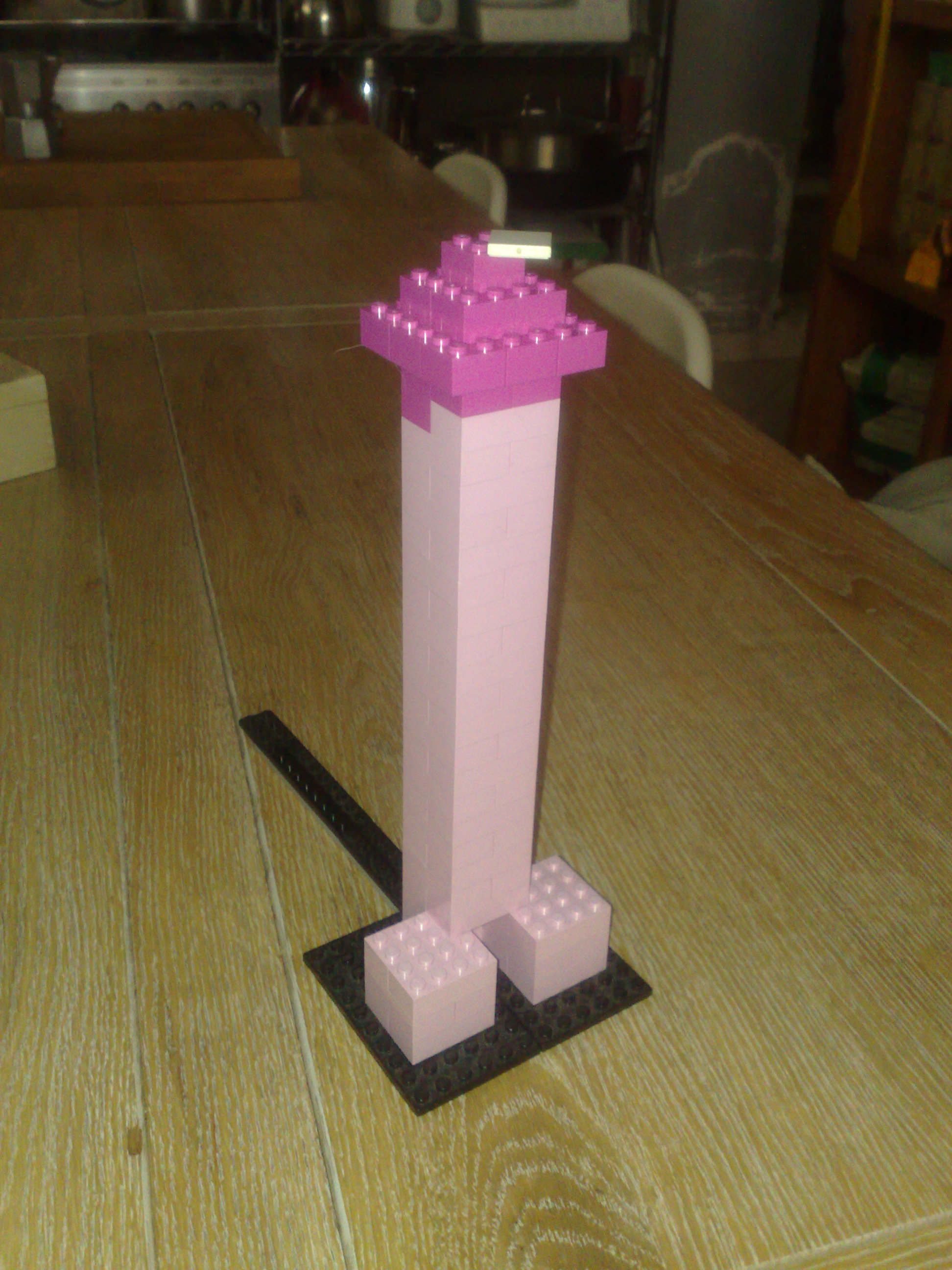 The image captures a creative scene set on a wooden dining table. The main subject of the image is a vibrant and unusual piece of tabletop artwork, taking the form of a tall, narrow tower-like structure. The structure is composed primarily of Lego bricks in shades of pink and white, with the pink pieces forming the outer shell and the white pieces filling the hollow interior. Adding an unconventional twist to the design, the base of the structure is a wide, stylized stand made entirely of Lego bricks, slightly offset to one side. This unique piece of Lego sculpture occupies a central position on the table, drawing the viewer's attention immediately. The background of the image, although partially obscured, suggests a lived-in space with various objects scattered around, providing a sense of context and scale to the Lego artwork. Overall, the image presents an intriguing blend of elements, with a focus on the playful interactivity between everyday objects and the world of Lego.