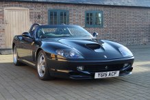 Pistonheads - The image shows a black sports car parked on a brick pavement in front of a building. The car features a prominent black hood and a rear spoiler, and it's oriented facing the camera. The vehicle has a license plate that reads "Y15 AFC". The building behind the car has visible brick cladding and what appears to be residential windows. The image captures the scene during daylight with ample natural light illuminating the car and the brick surface.