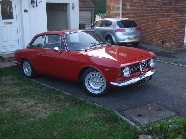 Let's see your Alfa Romeos! - Page 7 - Alfa Romeo, Fiat & Lancia - PistonHeads - This image displays a vibrant red classic sports car parked on a driveway adjacent to a white house with a chimney. The car is facing towards the left of the frame, and it appears well-maintained due to its shiny finish. In the background, another car is visible, parked on the street. The driveway itself has some grass sprouting on its edges, adding a touch of nature to the urban setting.