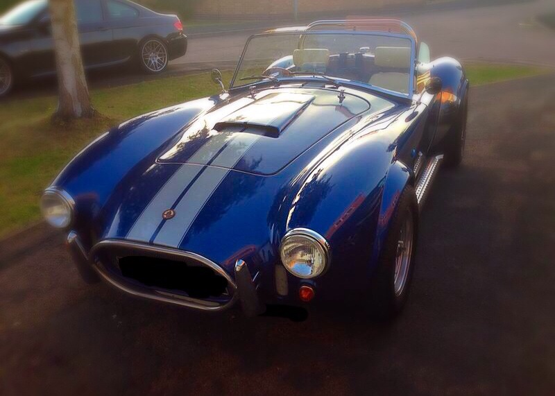 My life dream has come true! AC cobra time !! - Page 2 - Readers' Cars - PistonHeads - This image showcases a classic, blue convertible car parked on an asphalt surface. The car is sleekly designed with a shiny finish, and its hood is open, revealing a silver engine beneath. It's positioned under a clear sky with a blurred background that suggests trees and a distant building, creating a serene urban park setting. The car's door is closed, and there is a subtle reflection in its side that hints at the illumination of the scene.