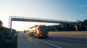 Pistonheads Switzerland Hgv - The image depicts a large transport truck traveling under an overpass on a significant highway. The truck is modern, with multiple sets of wheels and is in a state of brightness which might suggest either early morning or a sunset. The sky forms the backdrop of the image, exhibiting a gradient of colors that paints the scene with a soft, ethereal glow. Flanking the highway in the foreground is a line of stationary objects or structures that are partially obscured by the truck.