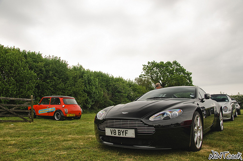 V8 Vantage - Page 1 - Middle East - PistonHeads - In this image, a black Aston Martin DB5 is the centerpiece, parked on a grass field with a silver sports car nearby. The cars are surrounded by lush green trees, creating a serene backdrop. A wooden fence can be seen in the background, adding a rustic charm to the scene. It appears to be an overcast day, dappling the surroundings with soft, diffused light. The image is marked with the text "Nate@Motive" in the bottom right corner, indicating the name of the photographer or designer.