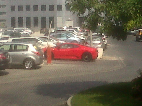 A car is stopped at a red light - Pistonheads - The image shows a street with multiple cars parked on the side. The focal point of the photo is a bright red sports car, which is parked diagonally across parking spots. Its color stands out among the other vehicles, which include a mix of sedans and SUVs in various colors. The vehicles are partially shaded by trees, and the street appears to be part of a residential or industrial area, suggested by the architecture of buildings in the background. The cars facing each other indicate the layout of parking spaces on this street.