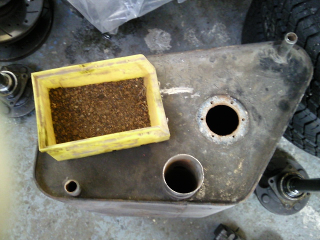 Anyone for a rust latte.. - The image shows an old, rusted utility box placed on its side in an indoor setting. It is a metallic box with two holes on its side and a yellow square attached to it. The box appears worn, possibly due to age or exposure to harsh conditions, and is situated on the floor alongside some other objects that are not the main focus of the image. Next to this box, there seems to be a hole in the floor or a part of a structure, and a tire or a circular object is visible in the background. The overall scene suggests a garage or a maintenance area with various equipment and parts.