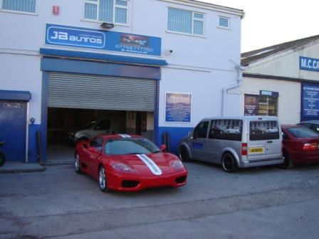 Autos Pistonheads Respray - The image features a large building with a high garage door. Parked in front of this door is a red sports car, and to the right, there is a gray van and a red car. On the building, the signboard reads " [Butos] Automotive" and "M.C Cars," indicating that this building is a car dealership or repair shop. The architecture suggests a traditional style. The area outside the building appears to be a busy parking lot with several other vehicles parked.