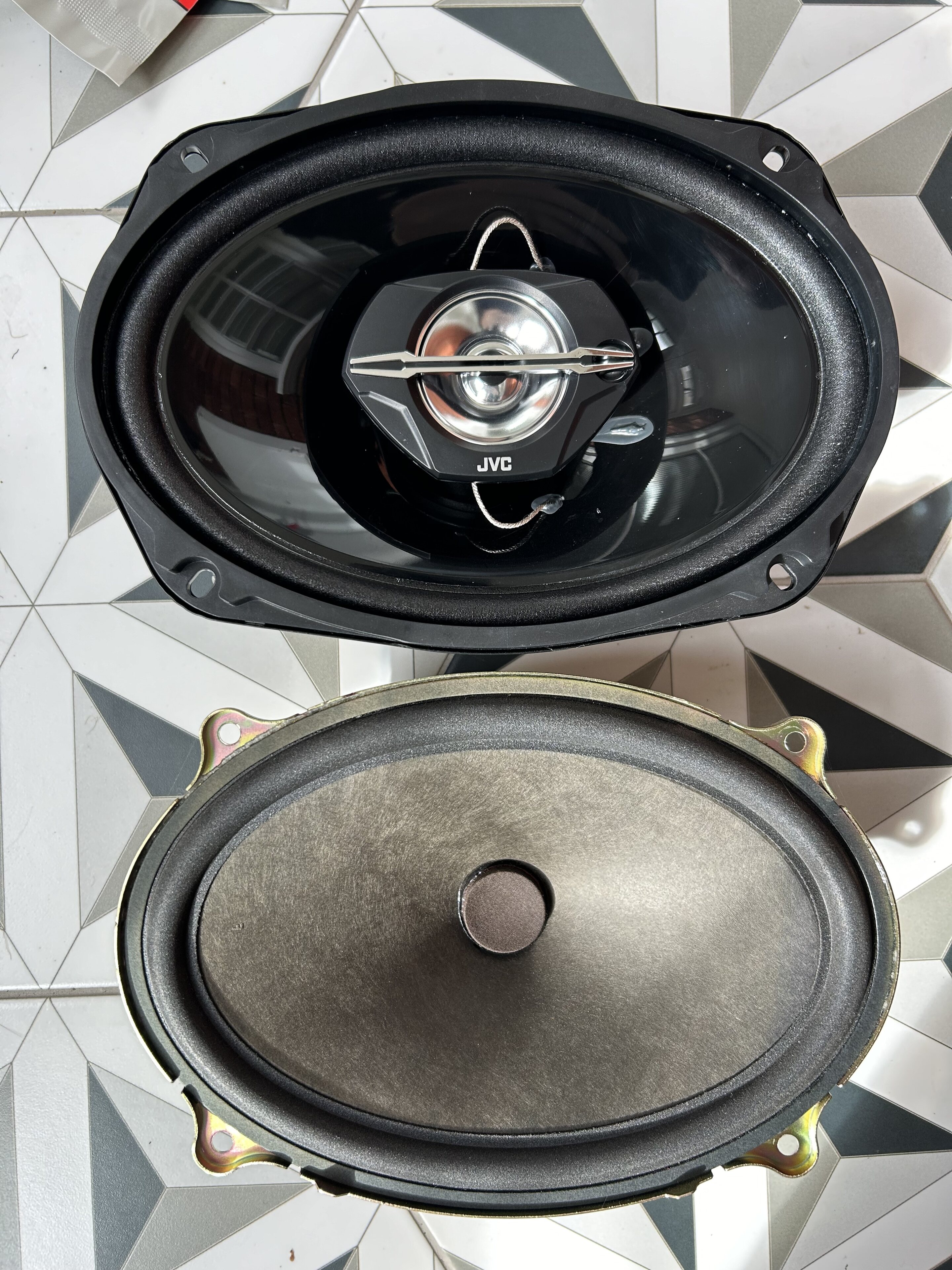 Pistonheads - The image shows two car speakers placed side by side against a white background. One speaker appears to be an older model with a black cone and silver trim, while the other is a newer model with a black cone and silver trim, but with some visible scratches on it. Both speakers are mounted in their respective metal frames, which have a shiny metallic finish. The photo seems to be taken from above to show the underside of the speakers and their mounting brackets. There is no text present in the image.