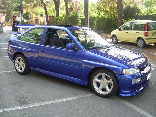 Burnt Pistonheads Memory - The image presents a vibrant city street scene with a focus on two cars parked adjacent to each other. The vehicle on the left is a striking blue sports car with a convertible top, exhibiting a sleek design and large, black rims. It's parked in a parking space marked by white lines. The vehicle on the right is a bright yellow car, also parked parallel to the blue car. The environment is urban, with buildings and a lush green park visible in the background. A pedestrian can be seen in the bottom left corner of the image. The day appears to be sunny, as indicated by the visibility and bright color of the cars.