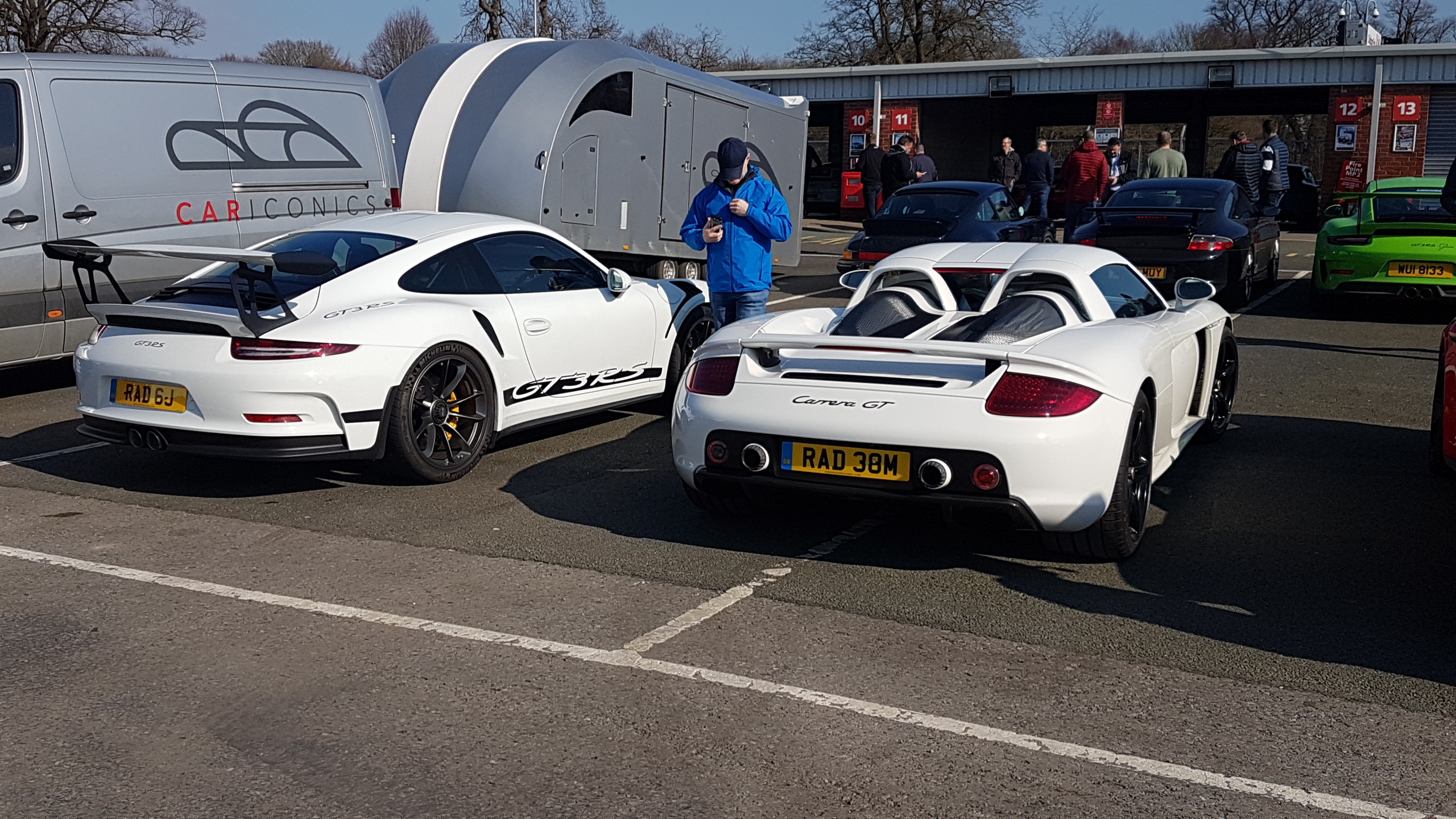 20 th rs track day. - Page 3 - 911/Carrera GT - PistonHeads - The image displays a scene in a parking lot where two high-performance sports cars are parked. The cars have distinctive designs, one featuring a prominent rear wing and the other with a smaller, flat spoiler on its trunk. They both have a sleek appearance with visible aerodynamic elements like side skirts and wide arches over the wheel wells.

There are two individuals present in the scene. One person is standing close to the car with the large rear wing, while the other individual is near the car with the smaller spoiler on its trunk. They appear to be engaged in some discussion or activity related to their vehicles.

The setting suggests a public parking area as there are other cars parked around them. There is no text visible within the image that provides additional context or information about the location or event. The lighting indicates it could be late afternoon or early evening, given the soft, diffused light on the scene.