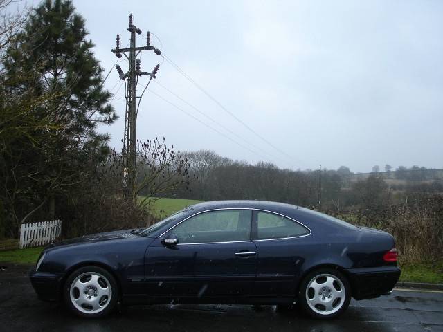 my V8 Merc - Page 1 - Readers' Cars - PistonHeads - The image features a dark blue car parked on a street, viewed from the front passenger side. The car is sleek, with a design indicative of European models. The sky is overcast, suggesting the photo was taken on a gloomy day. In the background, you can see an array of power lines and utility poles, along with leafless trees indicating that the photograph was likely taken in autumn or winter. The overall atmosphere of the image is somewhat somber and muted.