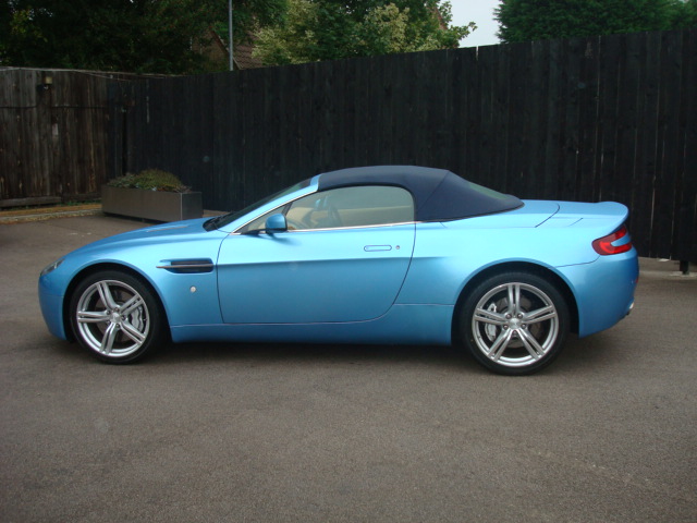 Drove a V8V Roadster - Page 1 - Aston Martin - PistonHeads - The image features a blue Aston Martin Vantage convertible sports car parked on a driveway. The car, which is the central focus of the image, is positioned against the backdrop of a wooden fence and a section of dark, leafy vegetation. The vehicle has a sleek design with a clean overall appearance. The driveway appears to be sloped and made of concrete, which complements the overall urban setting of the image.