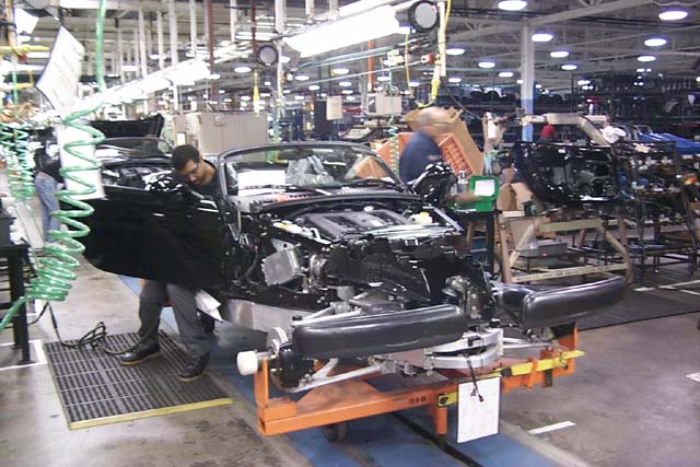 A group of people standing around a motorcycle - Pistonheads - This image depicts an indoor setting where the focus is on a car that appears to be in the middle of an assembly process or repair work. The car is being worked on within the confines of what looks like a factory or assembly plant. There are at least two people present in the scene; one of them is visibly inspecting the front of the vehicle, possibly ensuring its alignment. The floor is marked with blue and orange tape, suggesting an organized workspace where specific areas are designated for different tasks.