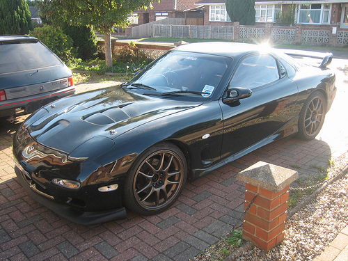 new member - Page 1 - East Anglia - PistonHeads - The image showcases a black sports car parked on a brick driveway, reflecting the sun in its polished frame. The car is equipped with aftermarket rear wings, adding a sporty aesthetic, and its sleek design is highlighted by a clean, glossy finish. It appears to be a two-door, with the driver's side visible, but there is no one seated in it. In the background, there are residential buildings, suggesting that this might be a suburban environment.