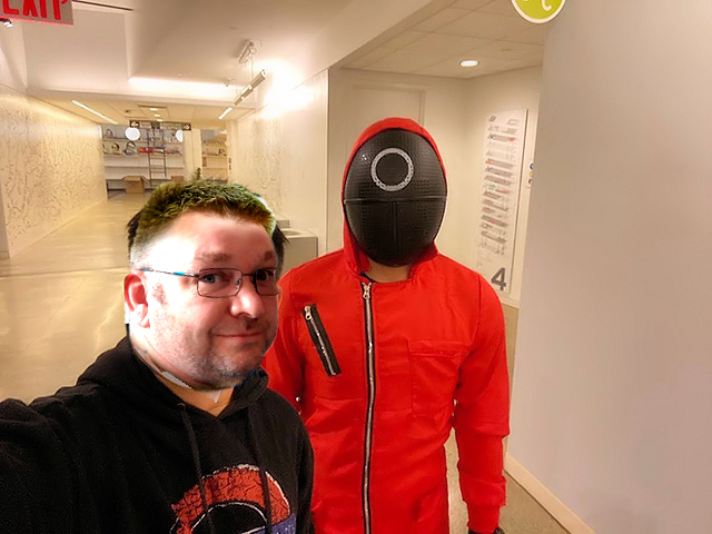 Photoshop Bobbers - Page 94 - The Lounge - PistonHeads UK - The image shows a person taking a selfie with another individual dressed in an unusual costume. The person taking the selfie is on the left, and they are wearing glasses and a black hoodie. To the right is another person wearing a red jacket over a hooded sweatshirt, paired with a mask. Both individuals are standing indoors, likely in an office or similar setting, as suggested by the office furniture visible in the background. The photo has been edited to add a humorous effect, with the individual on the right's image being altered to resemble a robot. The person taking the selfie appears to be laughing at this edit.