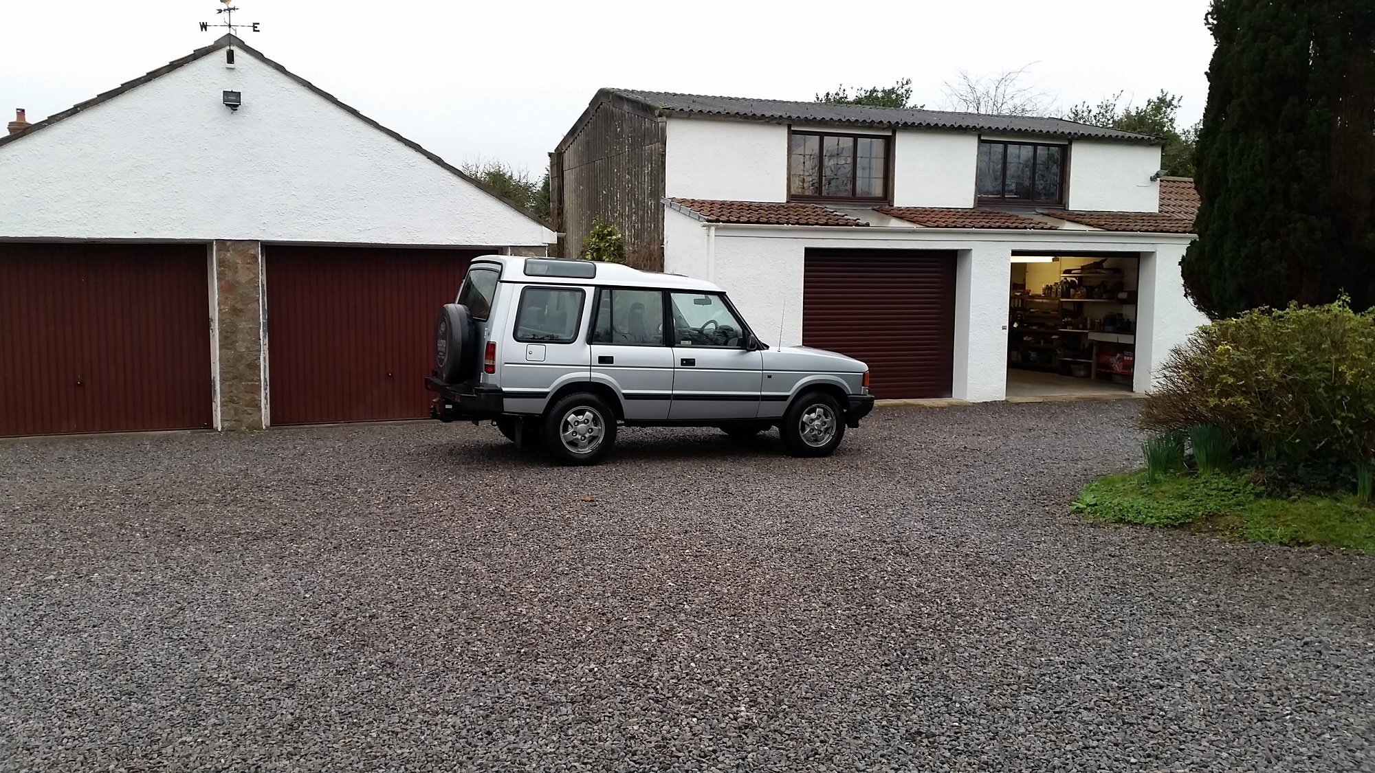 show us your land rover - Page 69 - Land Rover - PistonHeads - The image displays a gravel driveway leading to a house and a garage. At the end of the driveway is a silver SUV parked in front of the closed garage door. The house occupies the background, and the sky is overcast. The driveway is orderly, with gravel evenly spread. The image seems to depict a quiet residential area.