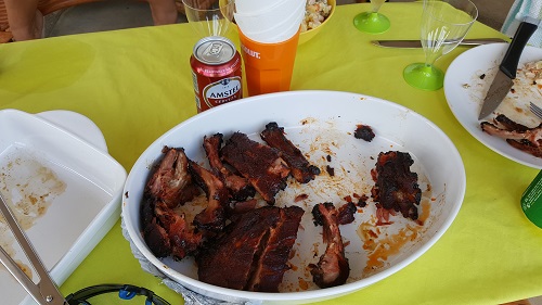 The bbq photo & recipe thread - Page 128 - Food, Drink & Restaurants - PistonHeads - The image presents a casual dining scene with a white bowl filled with barbecued ribs placed on a vibrant yellow tablecloth. In the background, a dining table is set with various utensils, including a knife, forks, and a couple of cups, one of which appears to contain a red beverage. The overall setting suggests a meal in progress, with the focus on the barbecued ribs in the front bowl. The lighting and outdoor decor hint at a backyard barbecue or outdoor gathering.