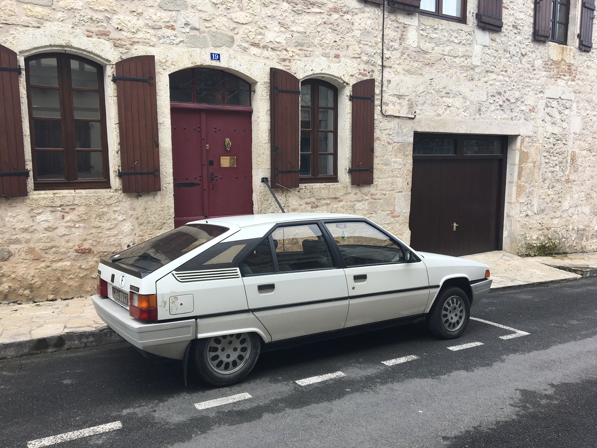 My bodged Citroen BX 16v - Page 16 - Readers' Cars - PistonHeads - The image features a white hatchback car parked in front of a building with a distinctive two-toned facade, blending beige and brown. The building is made of stone and has multiple arch windows. Beside the car, there is a red door, and to the right, a wooden garage door with a small black handle. The car is positioned in front of a white painted hatch, which is likely the entrance to the underground garage under the building.