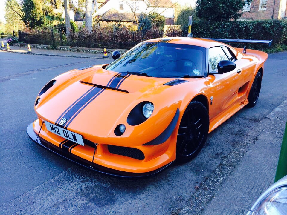 A red car is parked in a parking lot - Pistonheads - This image shows a vibrant, orange Lamborghini Huracan parked on the side of a lush green road. The car is sporting black tires with a diamond-spoke pattern and black trimming along the windows and stripes on the hood and sides. It has a sleek design with smooth curves and a low center of gravity. The frame captures a clear view of the front and side profile of the sports car, highlighting its luxury and performance status. There is a notable front lip spoiler and a rear wing, adding to the car's aerodynamic characteristics. The photo appears to be taken during the day under clear skies.