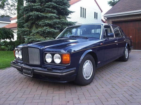 very clean looking 1989 Bentley Turbo R  - Page 1 - Rolls Royce & Bentley - PistonHeads - In the image, there is a classic blue Bentley car parked in front of a house. The Bentley, known for its luxury and elegance, is one of the most prestigious makes in the automotive industry. Its distinctive grille and round headlights are clearly visible. The car is parked on a brick driveway, and the house in the background has a red roof. There are trees in the vicinity as well. The automobile appears to be in good condition, indicating that it could be a rare or valuable model. Its classic design and color add to its appeal.