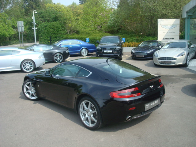 Gang Pistonheads - This image showcases a collection of modern cars parked in a lot. The primary focus is a sleek black sports car positioned in the foreground, which appears to be a luxury model with distinctive round headlights and distinctive red taillights. In the background, several other luxury cars are visible, including a mix of other sports cars and possibly some two-door coupes. The weather is clear, and the setting seems to be during the daytime. There's also another sports car on the far right side of the frame. The cars are parked in a manner that suggests the image might have been taken at a car dealership or exhibition.