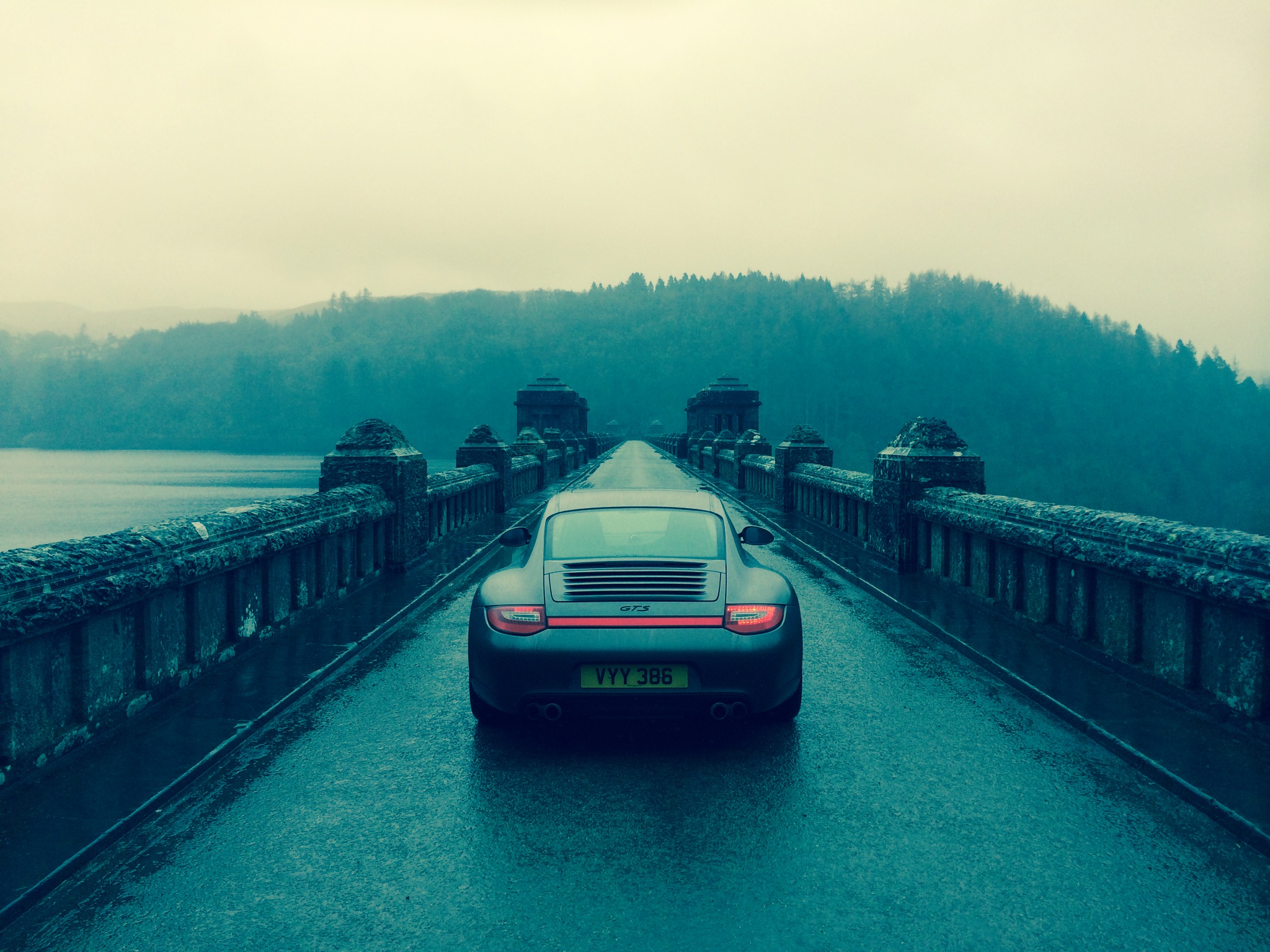 Show us your REAR END! - Page 219 - Readers' Cars - PistonHeads - The image captures a striking scene of a green and red sports car driving over a bridge with a dark sky as the background. The bridge appears to be in a mountainous area, with rolling hills sliding into the fog. The car is in motion, moving from the right side of the image towards the left, with the bridge stretching out ahead. The air is overcast, and the sky is gray, suggesting a cool or dreary day. The image conveys a sense of solitude and speed, as the car drives through the landscape.