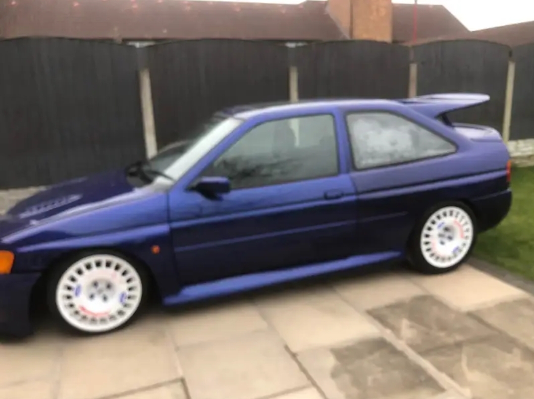 Pistonheads - The image shows a blue hatchback parked on a driveway. It has a custom body kit, including a wide-body design and fender modifications, which give it an aggressive and sporty appearance. The vehicle features white wheels with distinctive rims, contributing to its customized look. In the background, there is a building with a visible brick wall, partially obscured by the car. The photo seems to have been taken during the day under clear skies.